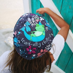 Puede incluir: Un sombrero de tela gris con estampado floral y una rana verde sentada en una luna creciente azul. El sombrero tiene el texto "toad-ally chilling" impreso.
