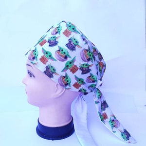 Puede incluir: Un gorro de quirófano blanco con una corbata. El gorro tiene un patrón repetido de un personaje de dibujos animados verde con orejas grandes.