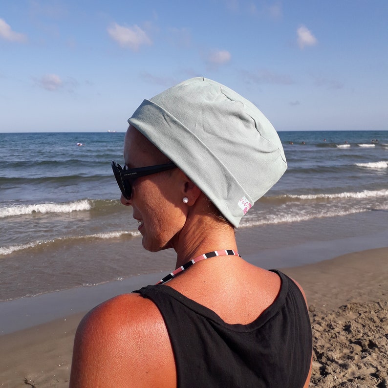 May include: A woman wearing a light blue beanie and black sunglasses stands on a sandy beach with the ocean in the background.