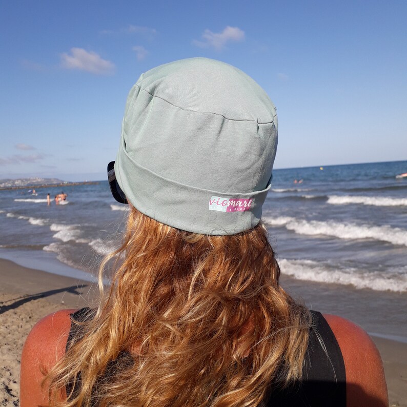 May include: A woman with long, wavy, light brown hair is wearing a light blue hat with the word "viomare" printed on it. She is standing on a beach with the ocean in the background.