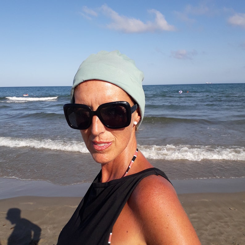 May include: A woman wearing a light blue beanie and black sunglasses stands on a sandy beach with the ocean in the background.