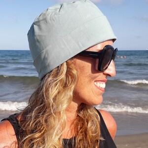 May include: A woman with long, wavy blonde hair is wearing a light blue cotton beanie and black sunglasses. She is smiling and looking to the right. The background is a beach with blue water and white waves.