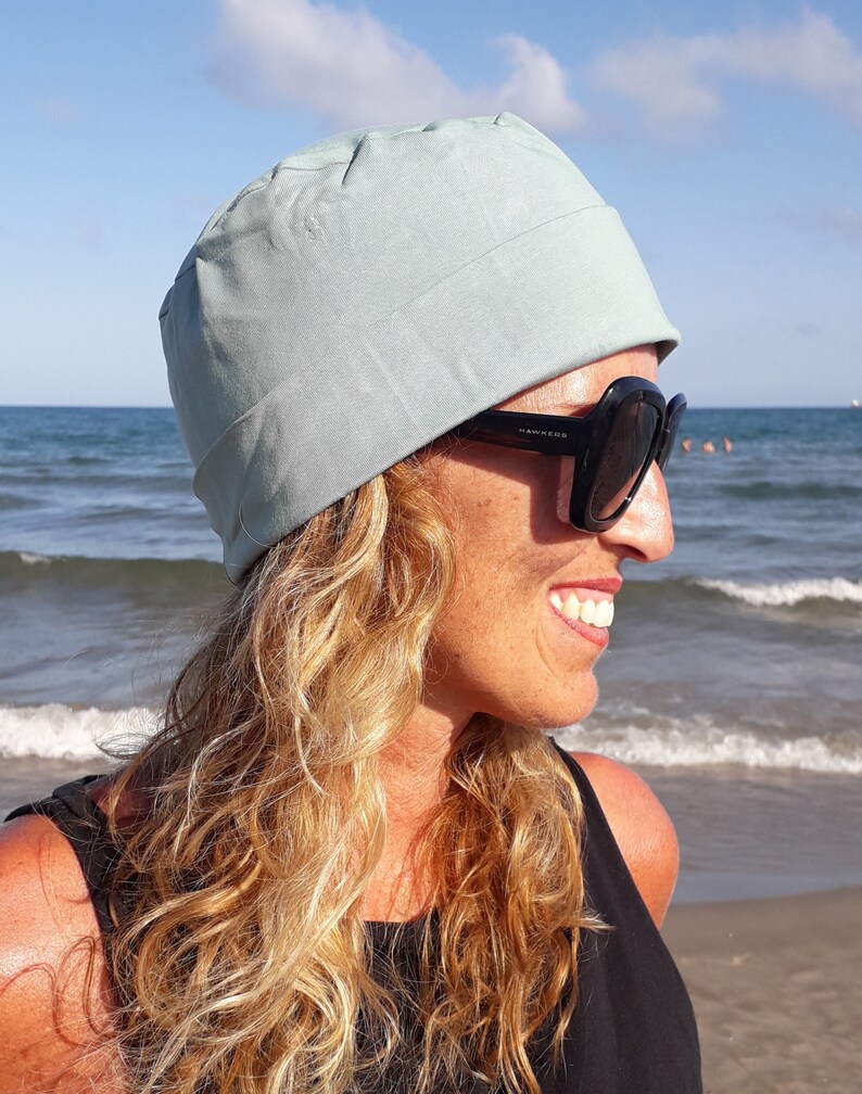 May include: A woman with long, wavy blonde hair is wearing a light blue cotton beanie and black sunglasses. She is smiling and looking to the right. The background is a beach with blue water and white waves.