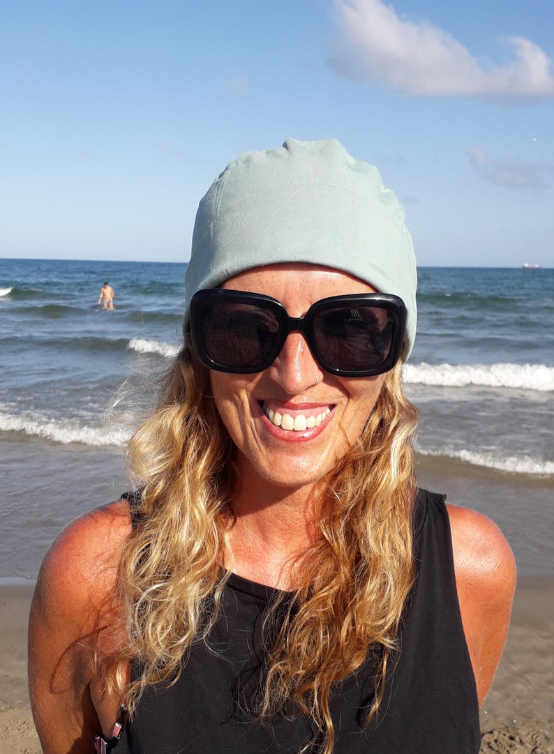May include: A woman with long blonde hair is wearing a light blue beanie and black sunglasses. She is smiling and standing in front of a beach with blue water and white waves.