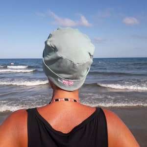 May include: A person wearing a light blue fabric hat with the word "remark" printed on it. The person is standing on a beach with the ocean in the background.