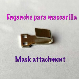 May include: A silver metal mask attachment with two holes on one end and a hook on the other end. The text "Enganche para mascarilla" and "Mask attachment" is visible in the image.