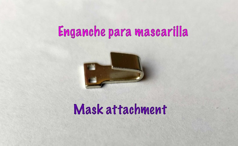 May include: A silver metal mask attachment with two holes on one end and a hook on the other end. The text "Enganche para mascarilla" and "Mask attachment" is visible in the image.