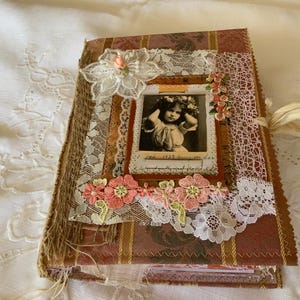 May include: A handcrafted journal with a burgundy and gold striped fabric cover, decorated with white lace, hessian, and pink floral appliqués. The centre features a vintage black and white photograph of a child with the handwritten text Jan 1922.