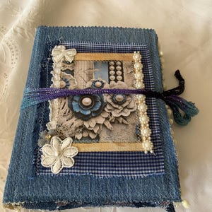 May include: A rectangular journal with a blue denim fabric cover. The front features a layered collage of blue gingham fabric, lace floral appliqués, a string of white faux pearls, and a central paper panel with blue flower illustrations tied with a purple ribbon.