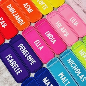 May include: A selection of brightly coloured popsicle sleeves, each bearing a different name in white lettering. The sleeves come in a variety of colours, such as red, orange, yellow, pink, purple, and blue. Names include "MIMA", "CHARLOTTE", "ASMI", "DYNJANDI", "HILARY", "LILY", "ELLA", "LYDIA", "PENELOPE", "ISABELLE", "WALT", "NICHOLAS", and "MATTHEW".
