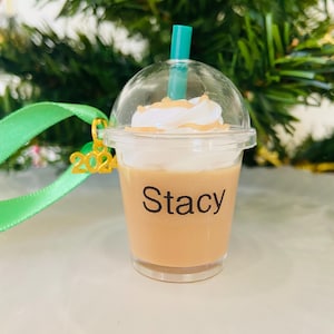 Puede incluir: Un vaso de pl&aacute;stico transparente con un l&iacute;quido marr&oacute;n y una cobertura de crema batida blanca. Una pajita verde sobresale de la parte superior. El vaso tiene un amuleto dorado 2022 unido a &eacute;l y una etiqueta negra que dice "Stacy".