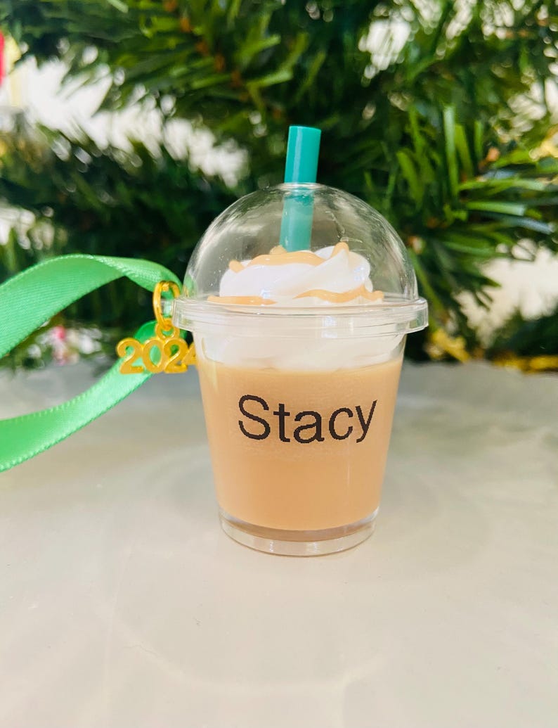 Puede incluir: Un vaso de pl&aacute;stico transparente con un l&iacute;quido marr&oacute;n y una cobertura de crema batida blanca. Una pajita verde sobresale de la parte superior. El vaso tiene un amuleto dorado 2022 unido a &eacute;l y una etiqueta negra que dice "Stacy".