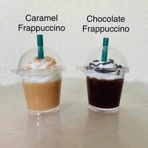 Puede incluir: Dos vasos de pl&aacute;stico en miniatura con tapas y pajitas, llenos de una bebida con sabor a caramelo y chocolate. Los vasos est&aacute;n etiquetados como "Caramel Frappuccino" y "Chocolate Frappuccino".