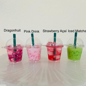Puede incluir: Cuatro vasos de pl&aacute;stico en miniatura llenos de l&iacute;quidos coloridos y cubitos de hielo. Los vasos est&aacute;n etiquetados como "Dragonfruit", "Pink Drink", "Strawberry A&ccedil;ai" y "Iced Matcha".