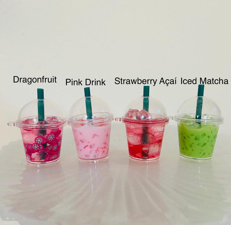 Puede incluir: Cuatro vasos de pl&aacute;stico en miniatura llenos de l&iacute;quidos coloridos y cubitos de hielo. Los vasos est&aacute;n etiquetados como "Dragonfruit", "Pink Drink", "Strawberry A&ccedil;ai" y "Iced Matcha".