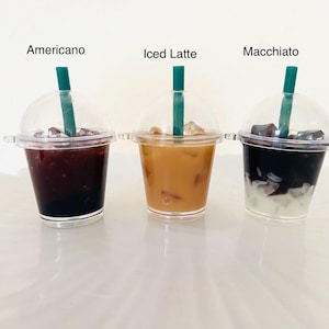 Puede incluir: Tres vasos de pl&aacute;stico transparente con tapas y pajitas llenos de diferentes bebidas de caf&eacute; helado. Los vasos est&aacute;n etiquetados como "Americano", "Iced Latte" y "Macchiato".