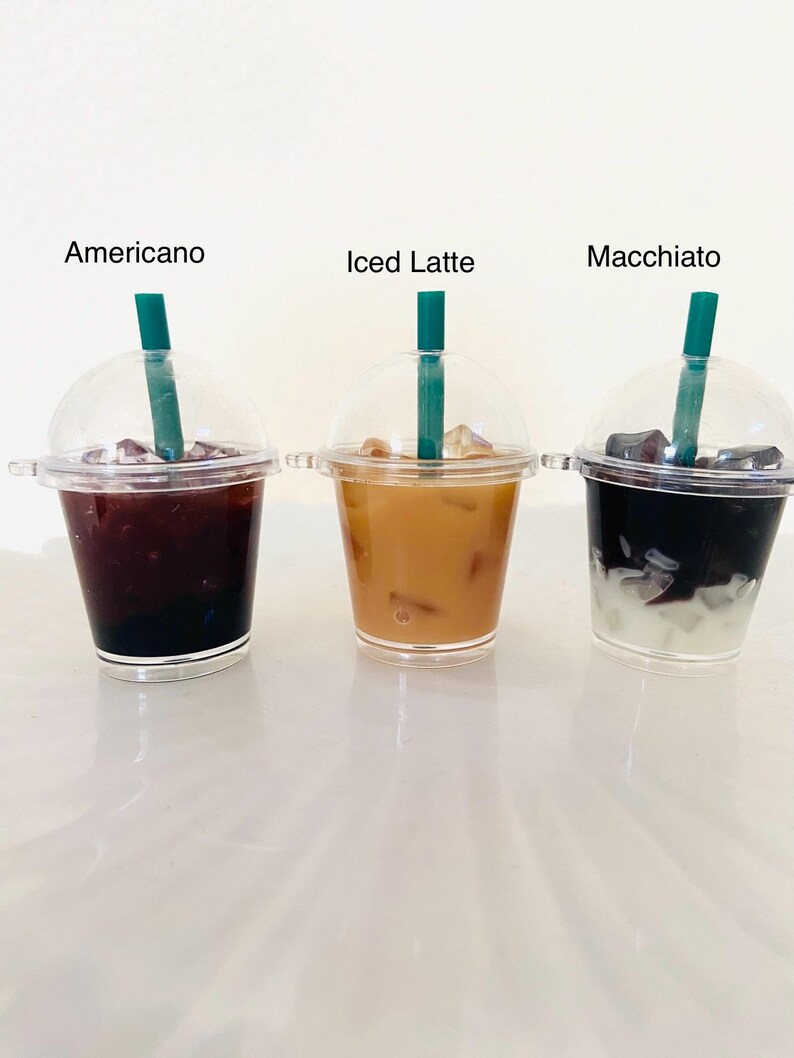 Puede incluir: Tres vasos de pl&aacute;stico transparente con tapas y pajitas llenos de diferentes bebidas de caf&eacute; helado. Los vasos est&aacute;n etiquetados como "Americano", "Iced Latte" y "Macchiato".