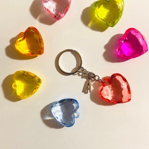 May include: A set of seven heart-shaped keychains in various colors, including pink, yellow, green, blue, and red. The keychains are made of clear plastic and have a faceted design. A silver keychain with a letter 'A' charm is included.