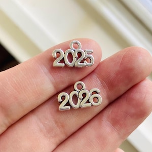 May include: Two silver-colored charms, one with the number 2025 and the other with 2026. Each charm has a small loop at the top for attaching to a necklace or bracelet. The charms are small and have a simple, elegant design.