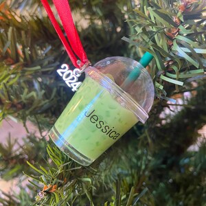 Puede incluir: Un adorno de pl&aacute;stico verde y transparente con forma de vaso de boba tea con una pajita. El adorno tiene el nombre "Jessica" escrito en &eacute;l y el a&ntilde;o "2024" en la parte superior.