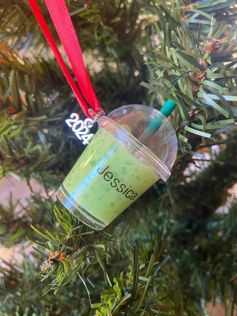 Puede incluir: Un adorno de pl&aacute;stico verde y transparente con forma de vaso de boba tea con una pajita. El adorno tiene el nombre "Jessica" escrito en &eacute;l y el a&ntilde;o "2024" en la parte superior.