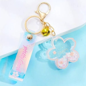 May include: A gold-colored keychain with a flower-shaped charm and a rectangular tag. The flower charm is light blue with pink accents and glitter. The tag reads "pretty good" in pink. A small gold bell hangs from the keychain.