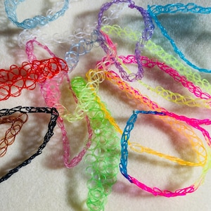 May include: A collection of colorful, stretchy tattoo choker necklaces in various shades including blue, red, pink, green, and purple. The necklaces are made of a transparent, plastic-like material and are arranged on a light-colored surface.