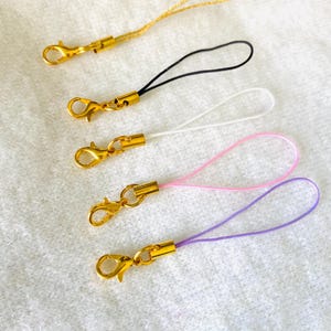 May include: Five colorful phone charms with gold-colored clasps. The charms feature loops in gold, black, white, pink, and purple. These accessories can be attached to phones, keychains, or other items.