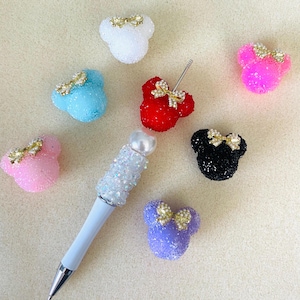 May include: A white pen with a textured, iridescent crystal-covered barrel and a pearl topper, surrounded by several mouse-shaped charms in white, light blue, pink, red, black, and purple, each featuring a small gold-tone bow accent.