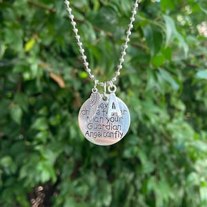 May include: Silver necklace with a charm that reads "Never fear more than your Guardian Angel can fly" and a letter A charm. The necklace is on a green leafy background.