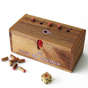 Puede incluir: Un juego de caja de madera llamado "Le Trou Du Cochon" con una cuerda roja y un dado. La caja tiene cinco agujeros con clavijas rosas y los números del 1 al 5. La caja tiene un círculo rosa con un cerdo dentro.