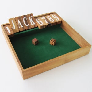 Può includere: Un tabellone da gioco in legno con una superficie di gioco in feltro verde. Il tabellone ha una cornice in legno e un set di tessere in legno con le lettere "JACK" e i numeri "1, 6, 7, 8, 9" su di esse. Due dadi marroni con punti neri sono sulla superficie in feltro.