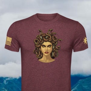 Puede incluir: Una camiseta de color granate con un gr&aacute;fico de Medusa, una figura mitol&oacute;gica griega con serpientes como cabello. El gr&aacute;fico est&aacute; en tonos de verde, marr&oacute;n y dorado. La camiseta tiene una peque&ntilde;a bandera estadounidense en la manga izquierda y una estrella en la manga derecha.