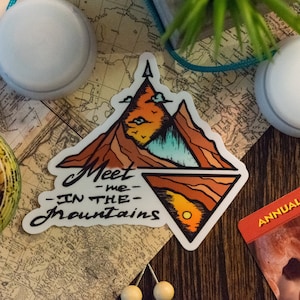Könnte beinhalten: Ein weißer Aufkleber mit einer Berglandschaft in Orange-, Braun- und Blautönen. Der Text "Meet me in the mountains" ist in schwarzer Kursivschrift geschrieben.