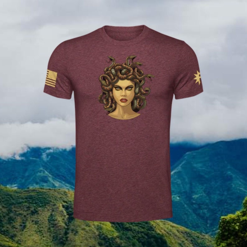 Puede incluir: Una camiseta de color granate con un gr&aacute;fico de Medusa, una figura mitol&oacute;gica griega con serpientes como cabello. El gr&aacute;fico est&aacute; en tonos de verde, marr&oacute;n y dorado. La camiseta tiene una peque&ntilde;a bandera estadounidense en la manga izquierda y una estrella en la manga derecha.