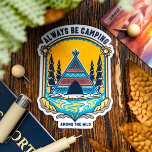 Può includere: Un adesivo con un design giallo, blu e verde. Presenta una tenda con un lago e alberi. Il testo recita "Always Be Camping Among The Wild".