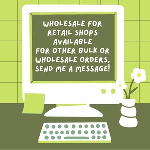 May include: A computer screen with a green background and a white border displays a message: "WHOLESALE FOR RETAIL SHOPS AVAILABLE FOR OTHER BULK OR WHOLESALE ORDERS, SEND ME A MESSAGE!" A green sticky note is on the computer screen. A keyboard is in front of the computer. A white vase with a green and white pattern holds two white flowers with yellow centers.