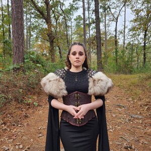 May include: A woman wearing a black dress with a brown leather corset and a white fur shawl. She is standing in a forest with trees in the background.