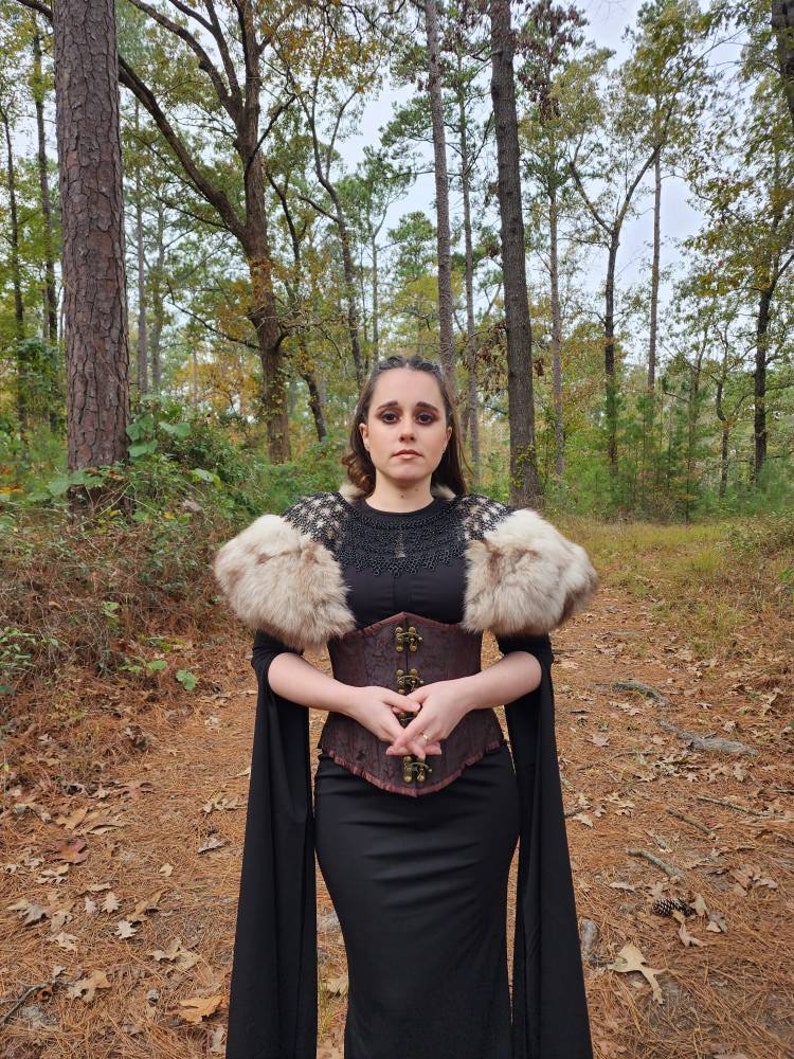 May include: A woman wearing a black dress with a brown leather corset and a white fur shawl. She is standing in a forest with trees in the background.