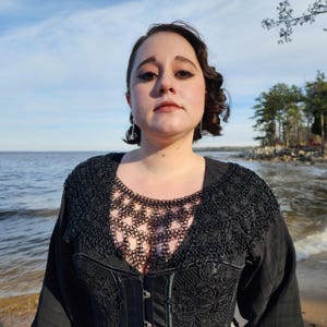 May include: A woman wearing a black top with a chainmail collar and a black lace overlay. She is standing in front of a body of water with trees in the background.