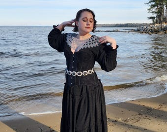 May include: A woman wearing a black dress with a lace neckline and a silver belt with rhinestones. She is standing on a sandy beach with water in the background.