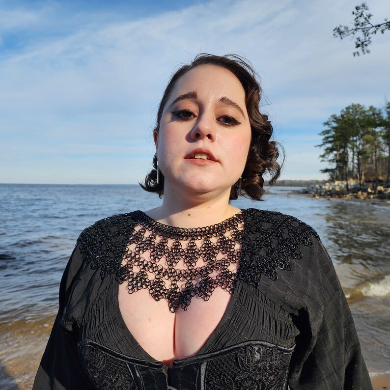May include: A woman wearing a black lace top with a black chainmail necklace. She is standing in front of a body of water with a shoreline in the background.