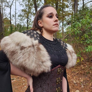 May include: A woman wearing a brown corset with metal clasps, a black top, and a fur collar. She is standing in a wooded area with fall leaves on the ground.