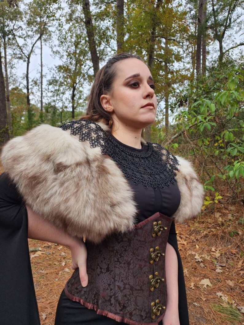 May include: A woman wearing a brown corset with metal clasps, a black top, and a fur collar. She is standing in a wooded area with fall leaves on the ground.