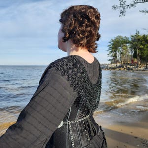 May include: A person wearing a black corset with a black chain and a black chainmail collar. The person is standing on a beach with the ocean in the background.