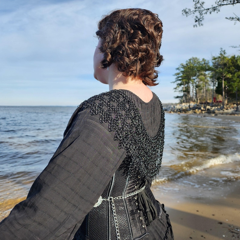 May include: A person wearing a black corset with a black chain and a black chainmail collar. The person is standing on a beach with the ocean in the background.