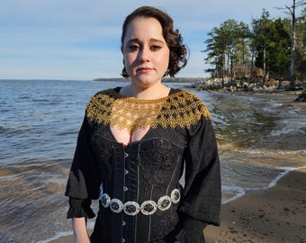 May include: A woman wearing a black dress with a gold chainmail collar and a silver and black belt. She is standing on a sandy beach with water in the background.