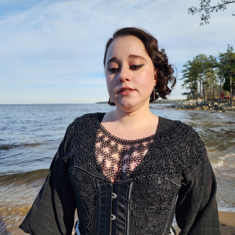 May include: A woman wearing a black lace top with a chainmail collar. She is standing on a beach with the ocean in the background.