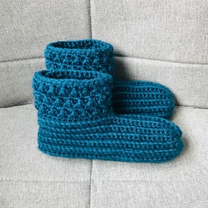 May include: A pair of teal blue crocheted slipper socks. The socks are made with a textured stitch and have a folded cuff.