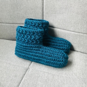 K&ouml;nnte beinhalten: Ein Paar tealblaue geh&auml;kelte Hausschuhe. Die Hausschuhe sind mit einem strukturierten Stich gearbeitet und haben ein weiches, gem&uuml;tliches Aussehen.